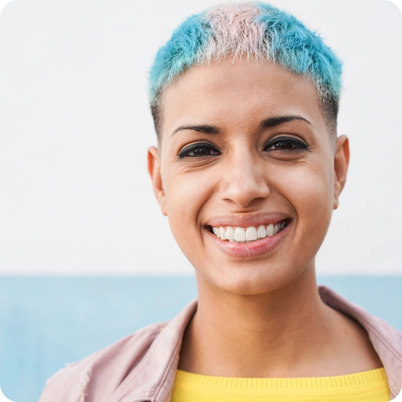 A confident smiling person with a short buzz cut hairstyle dyed turquoise blue fading to pink, wearing eyeliner, a light pink jacket over a yellow top, posed against a soft blue background. This image embodies the empowerment and respect gained through support for transgender and gender diverse individuals.