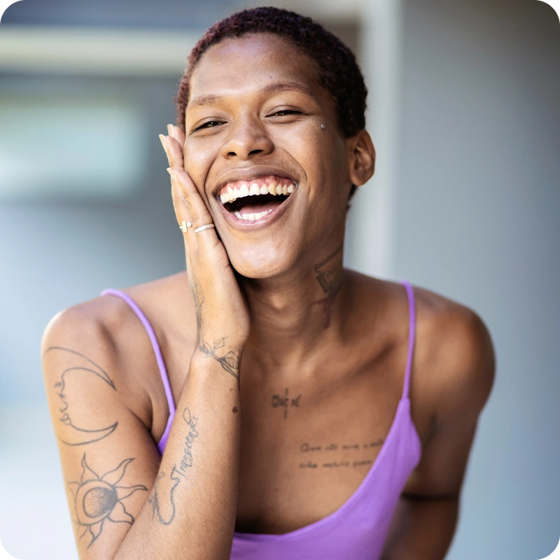 A joyful person with short curly hair, a facial piercing, and visible tattoos on their arms and chest, wearing a lavender tank top, smiling widely with hand resting on their cheek.