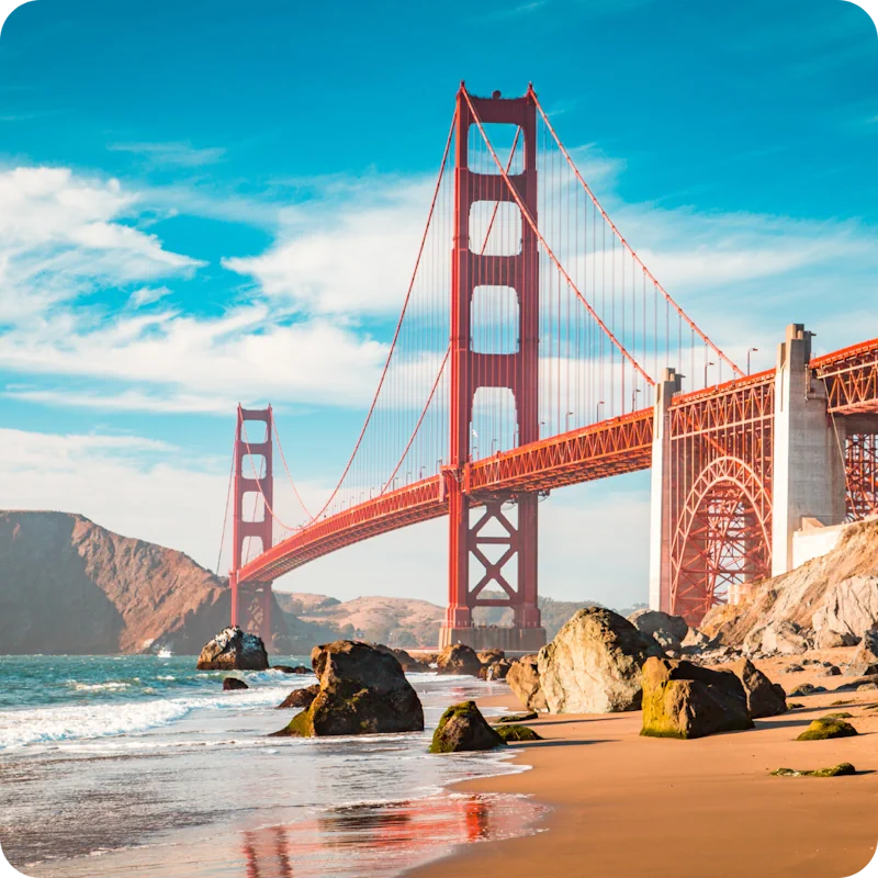 Iconic Golden Gate Bridge in San Francisco, viewed from a rocky beach with gentle waves lapping at the shore, under a bright blue sky with scattered clouds.