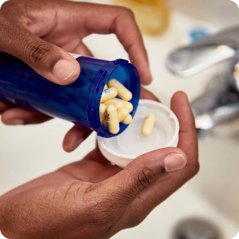 Close-up of hands pouring yellow capsules from a blue prescription bottle into the white cap, with a bathroom sink and faucet in the background.