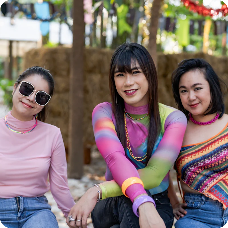 Three individuals sitting together, wearing colorful and vibrant clothing with accessories, smiling and showing support, set against a festive outdoor background with straw bales and greenery, reflecting the unique and authentic journeys of patients at ART Surgical.