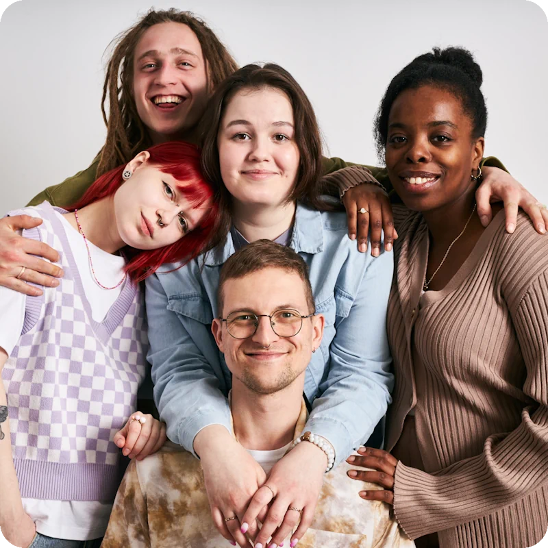 A group of five diverse individuals with colorful clothing and hairstyles, including a person with red hair and a checkered sweater, embrace each other warmly, symbolizing the importance of supportive communities for transgender mental health.