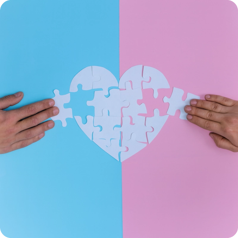 Two hands assembling a heart-shaped puzzle across a split blue and pink background, symbolizing the connection and support provided by mental health resources for transgender individuals.