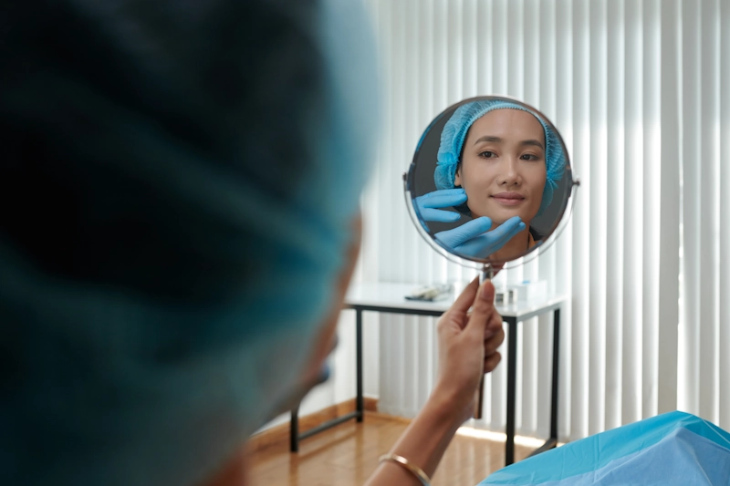 A person wearing a surgical cap and gown holds a mirror, calmly examining their face as a medical professional gently touches their chin, symbolizing reflection and care during a gender-affirming revision surgery consultation.