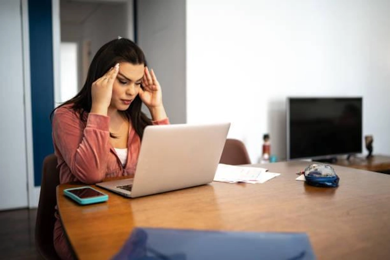 A stressed individual rubbing their temples while sitting at a laptop, representing how stress affects hormones and healing during a gender transition.