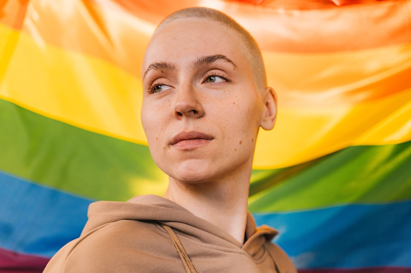 A confident nonbinary individual looking forward with a rainbow Pride flag in the background, representing empowering outcomes in gender-affirming nonbinary surgery.