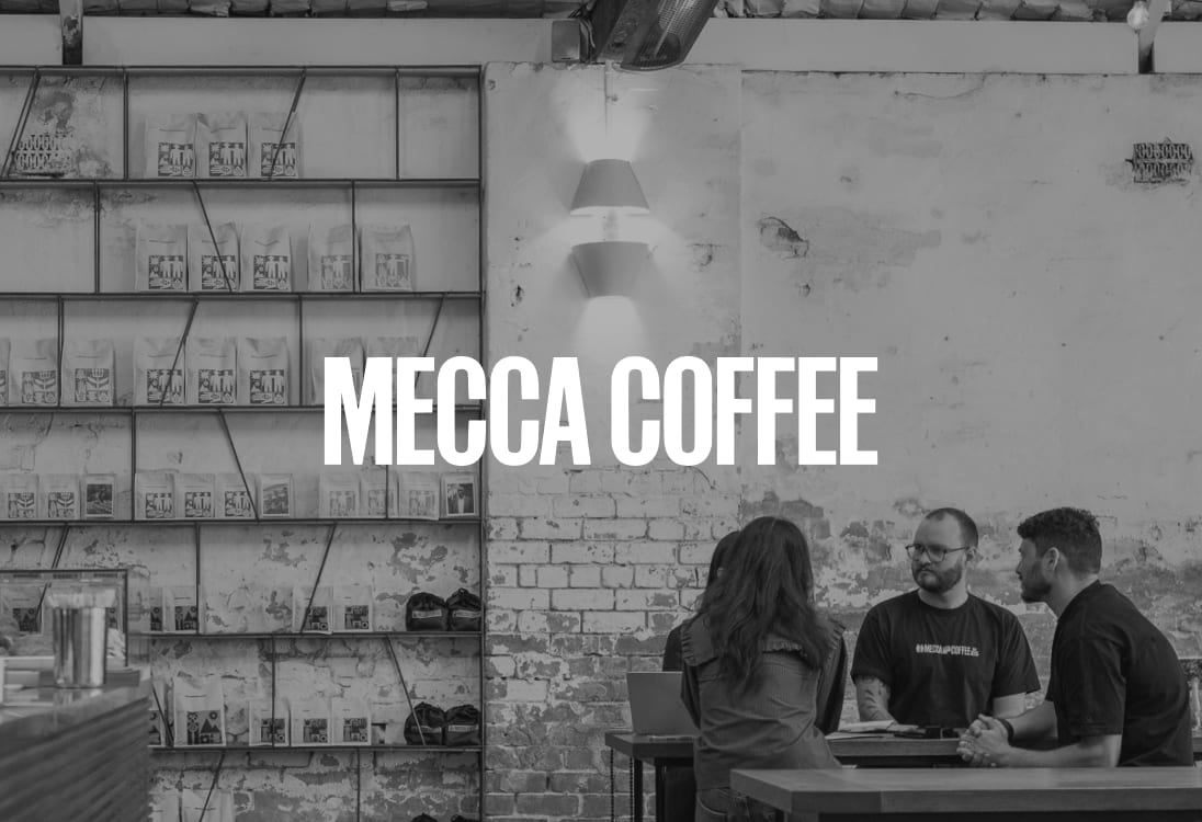 People sitting at a table in a rustic Mecca Coffee cafe, with shelves of coffee bags on the wall