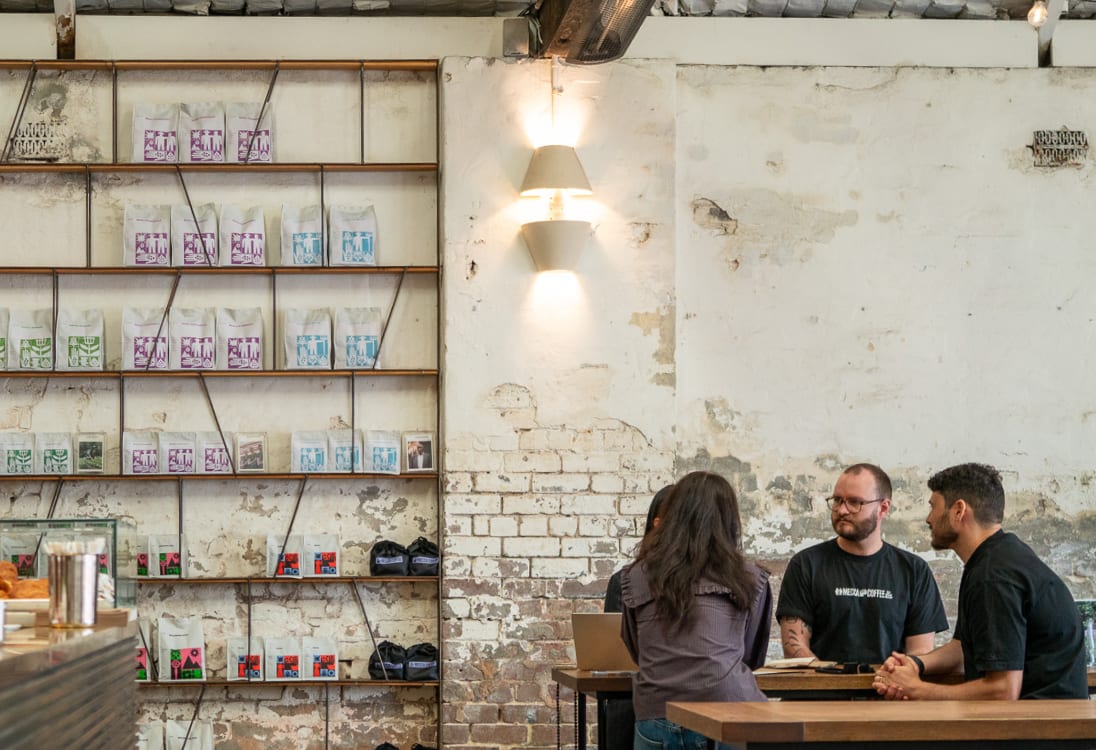 Four people seated at a wooden table in a rustic coffee shop with shelves of coffee bags on a brick wall.