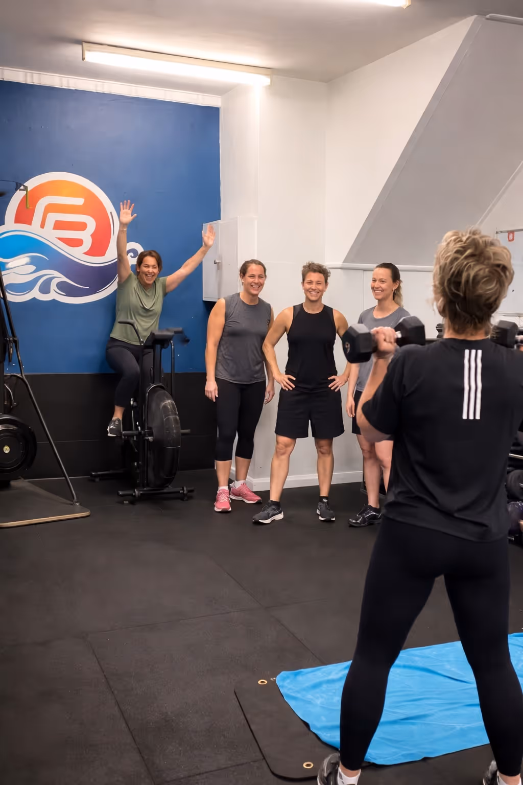 Four women in workout clothes smiling and watching another woman lifting dumbbells at Coastal Bodies gym.
