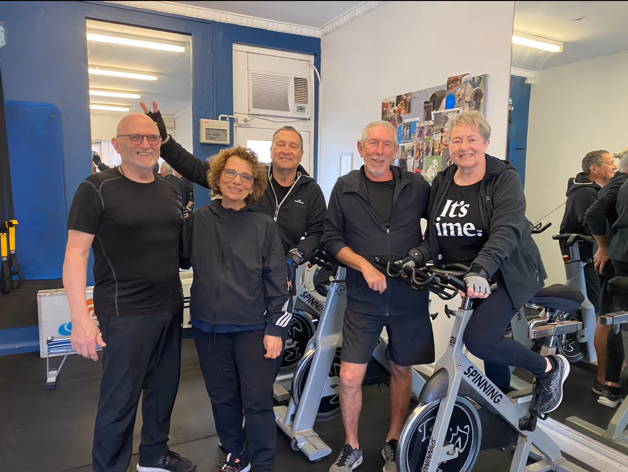 Coastal Bodies trainer Steve with group of four adults smiling at the gym and one woman sitting on a stationary spinning bike.