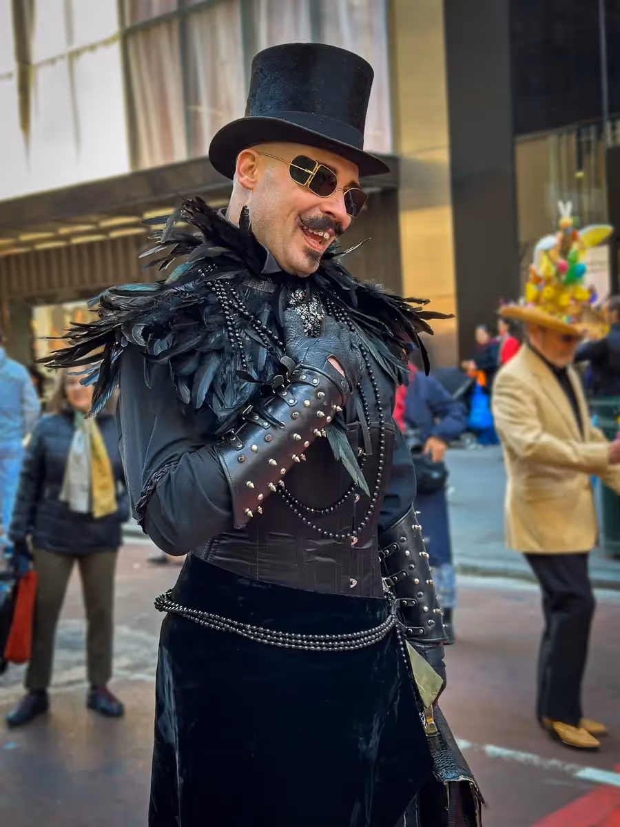 2024 Easter Bonnet Parade, NYC: Man in black jacket with studded cuff, feather shawl , black tophat and gasses