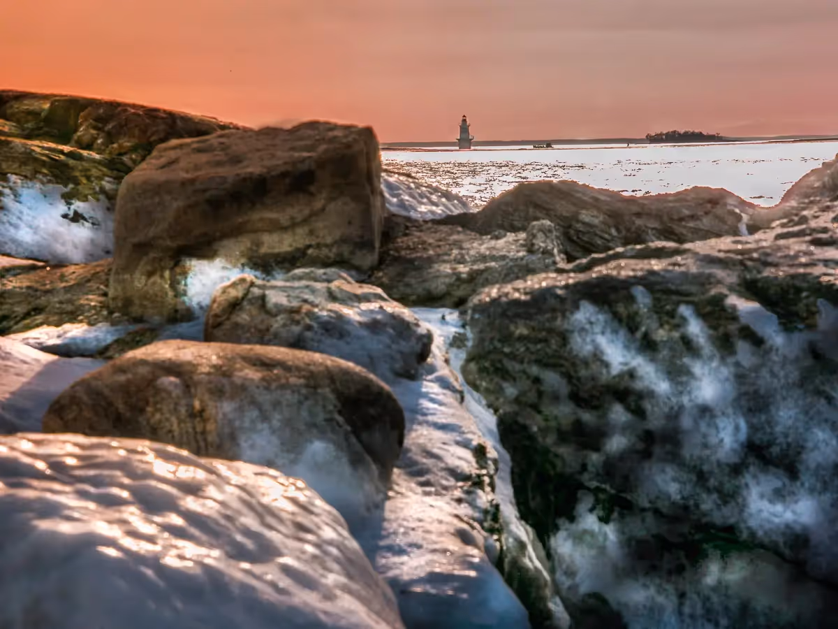You see a lighthouse in the distance over icy covered rocks in the jetty at sunset