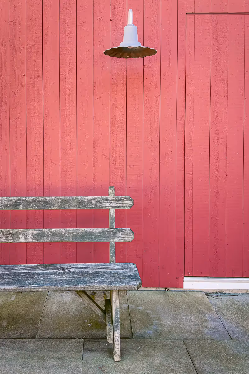 Simple yet inviting bench against a  wall of a red painted wooden home and a single fixture hanging just above the bench