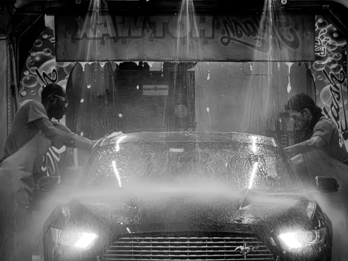Black and white Mustang going through a car wash with 2 people scrubbing and the water showering down