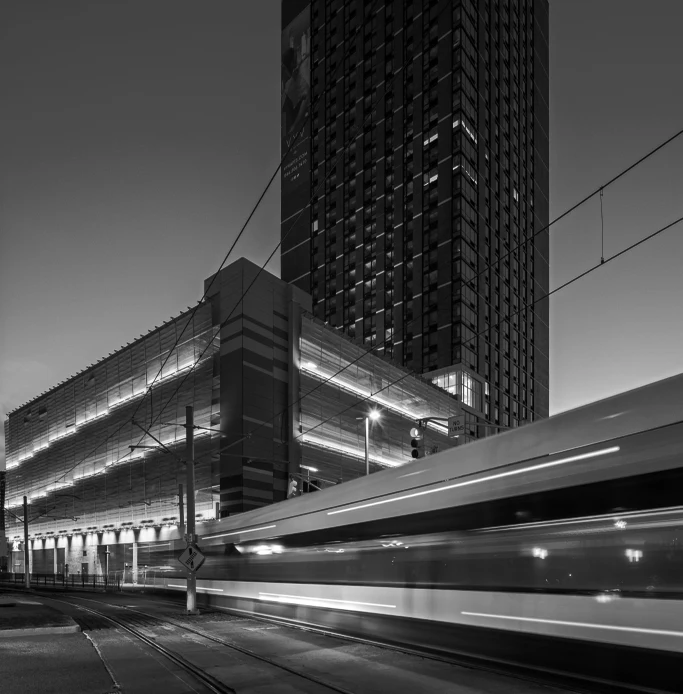 Black and white photo of a tower and parking structure at dusk with a passing light rail train