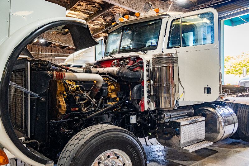 White semi-truck inside repair shop with hood open, exposing Caterpillar diesel engine, radiator, hoses, and exhaust system for maintenance inspection.
