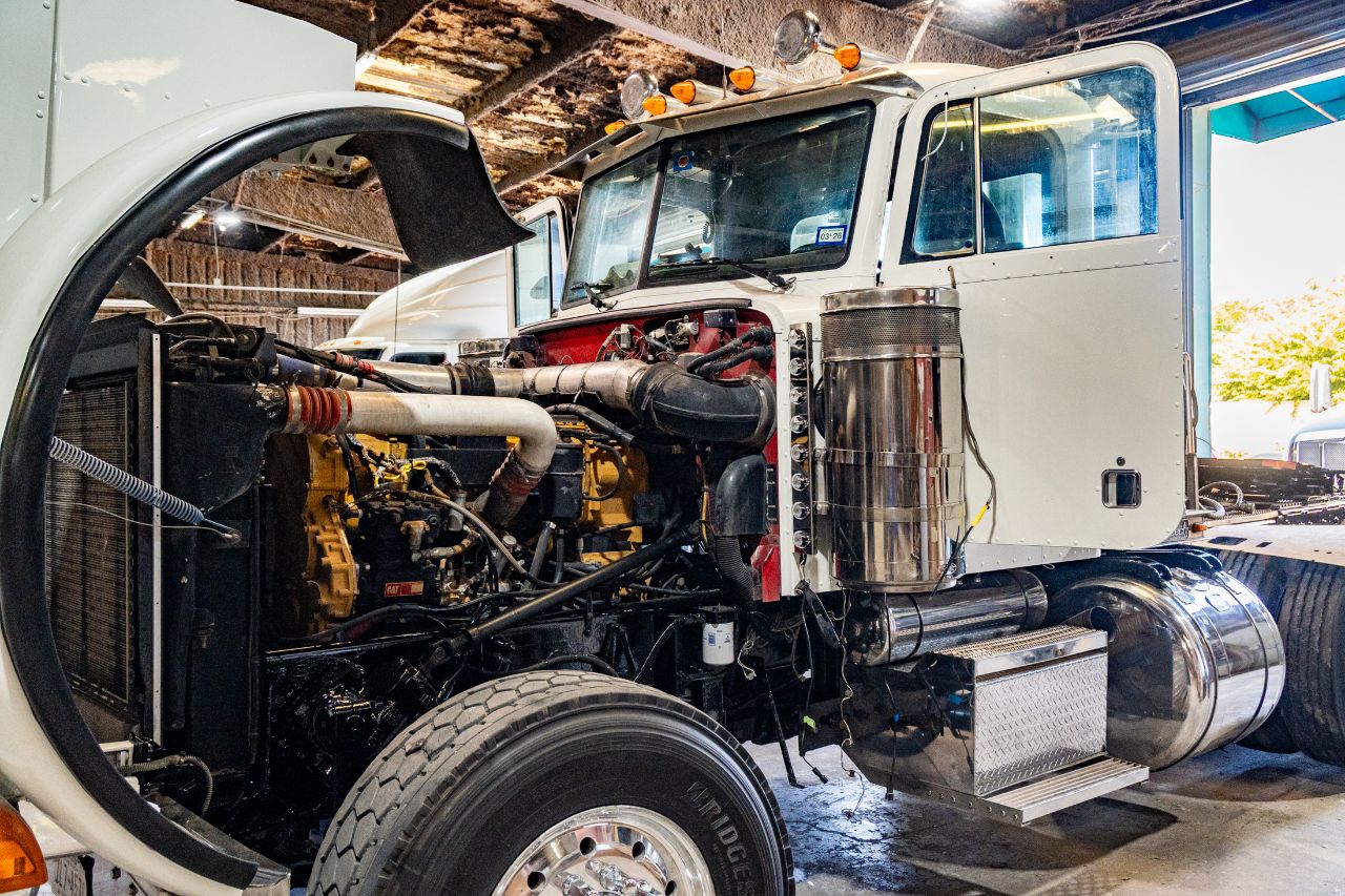 White semi-truck inside repair shop with hood open, exposing Caterpillar diesel engine, radiator, hoses, and exhaust system for maintenance inspection.