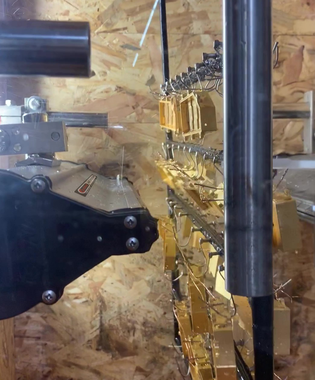 A close-up photograph capturing an active electrostatic powder coating process in an industrial spray booth with an OSB (oriented strand board) textured wooden background. A black automated powder coating gun, showing metal fittings and a section of a label ending in "AR SYSTEM", is mounted on the left and actively emitting a fine stream of powder. Suspended on a complex metal hook conveyor system on the right are dozens of intricately shaped golden-yellow metal components, arranged in multiple tiers and passing in front of the gun's spray path. The entire scene is framed by large metal cylindrical structures. The image focuses on the precise application of the coating.