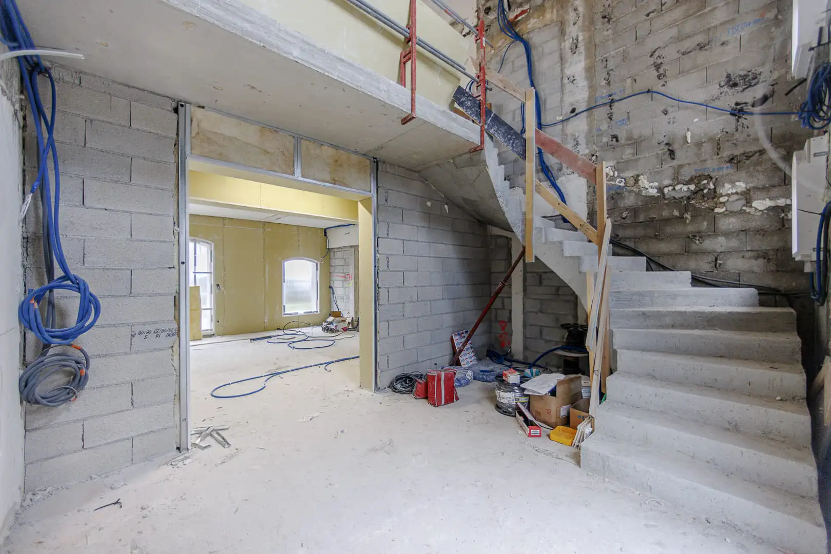 Intérieur de villa en cours de rénovation avec escalier en béton brut, murs en maçonnerie apparente et réseaux électriques en attente.