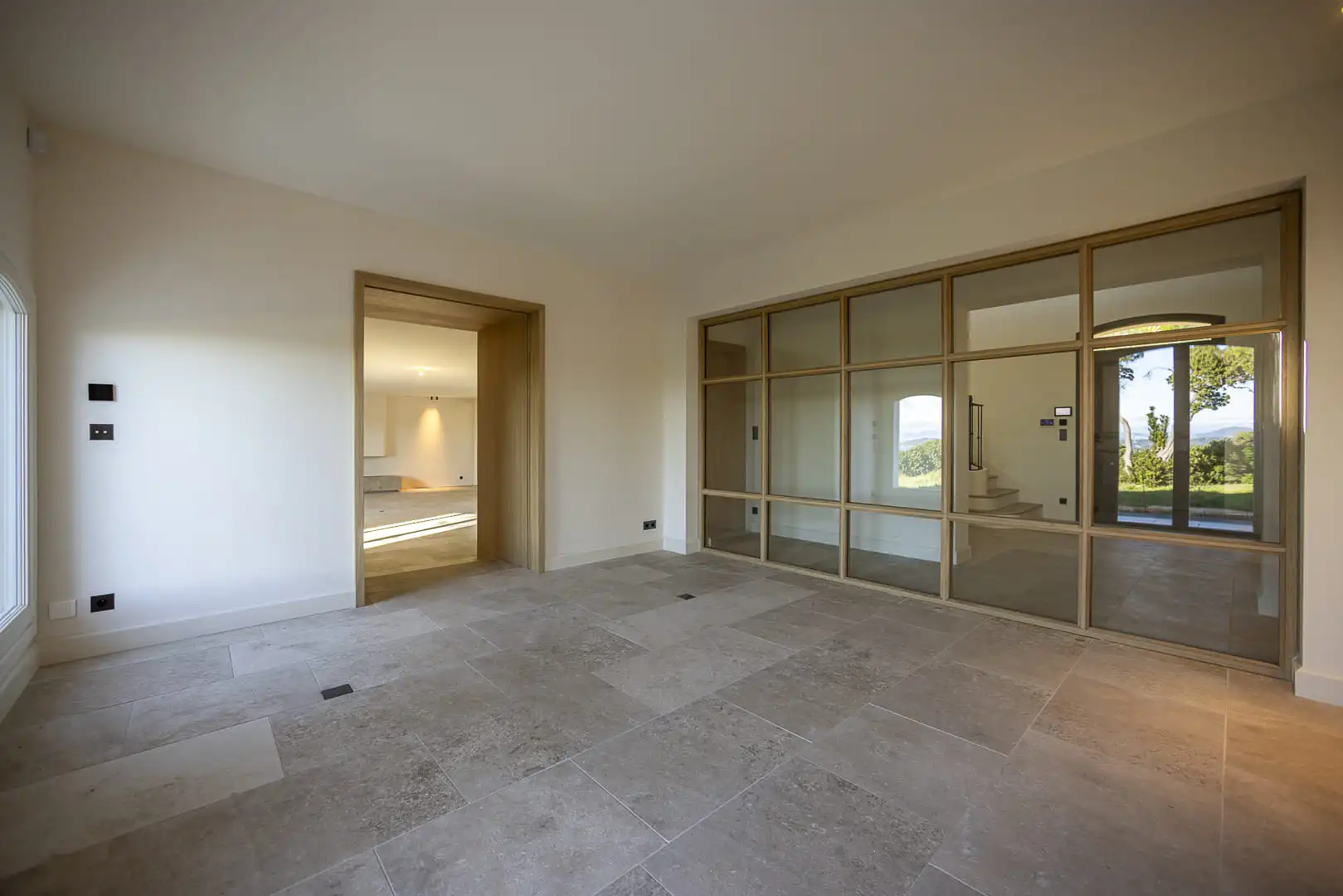 Espace intérieur finalisé avec verrière en menuiserie bois, sol en pierre naturelle et ouverture visuelle vers le paysage.