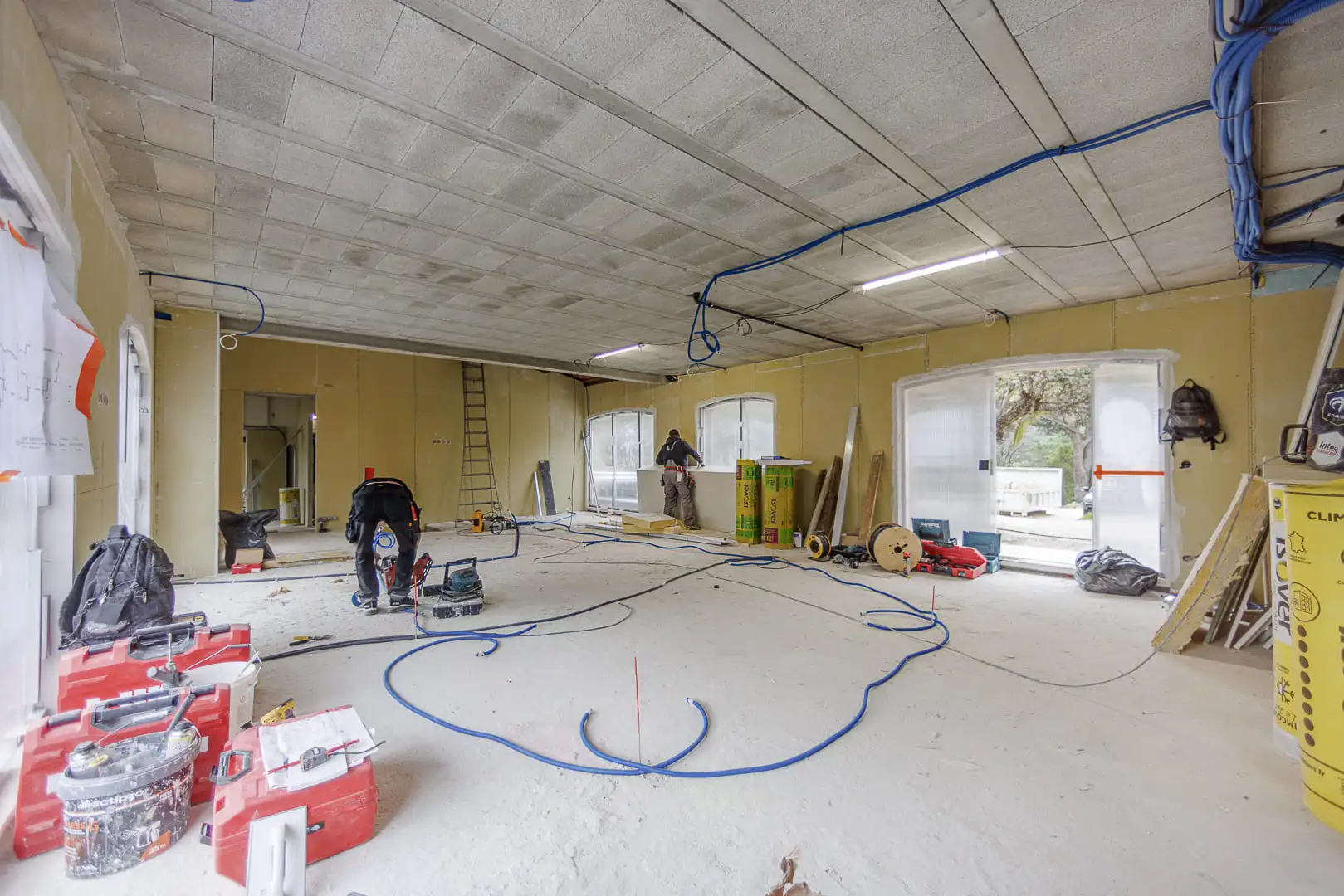 Grand espace de vie en rénovation avec intervention des équipes, réseaux techniques au plafond et larges ouvertures vers l’extérieur.