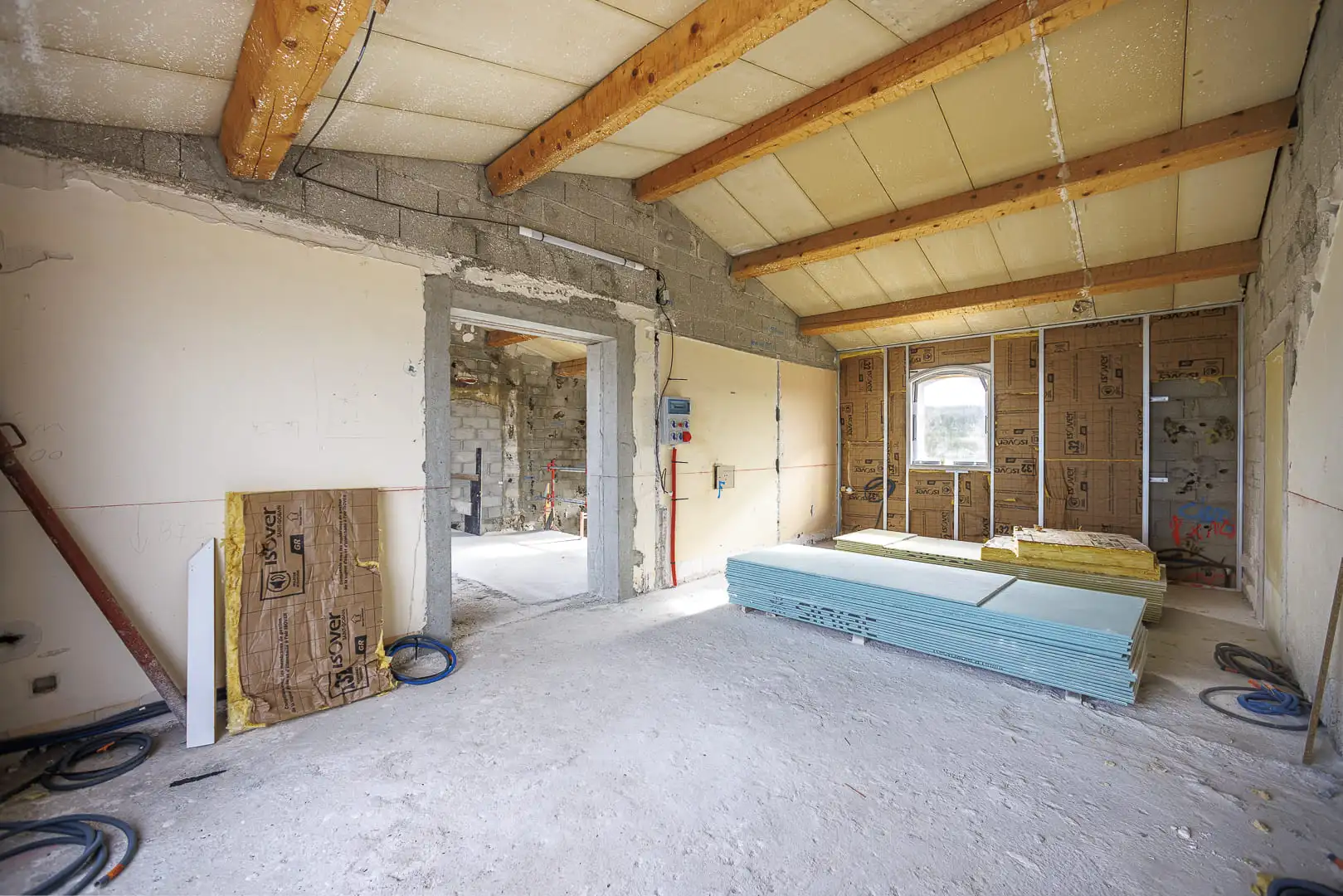 Espace en rénovation avec poutres en bois apparentes, isolation en cours de pose et plaques de plâtre stockées au sol.