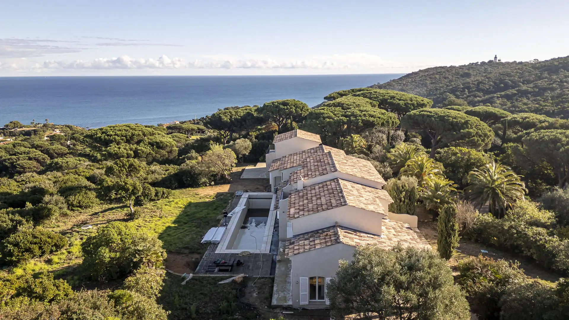Vue aérienne d’une villa rénovée entourée de pins parasols et palmiers, avec piscine extérieure et panorama sur la mer Méditerranée.
