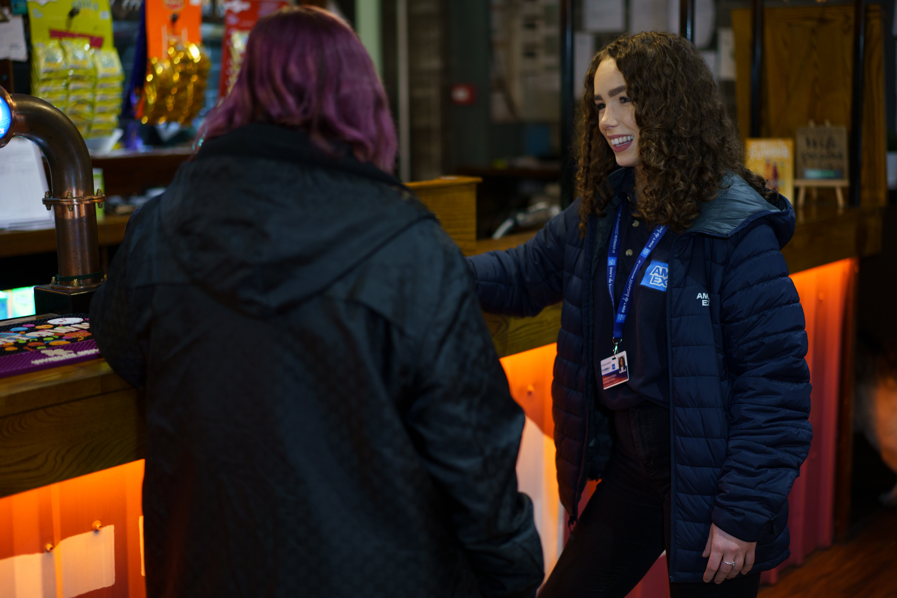 Smiling woman with curly hair wearing a navy blue American Express jacket and lanyard, talking to a person with purple hair at a counter.