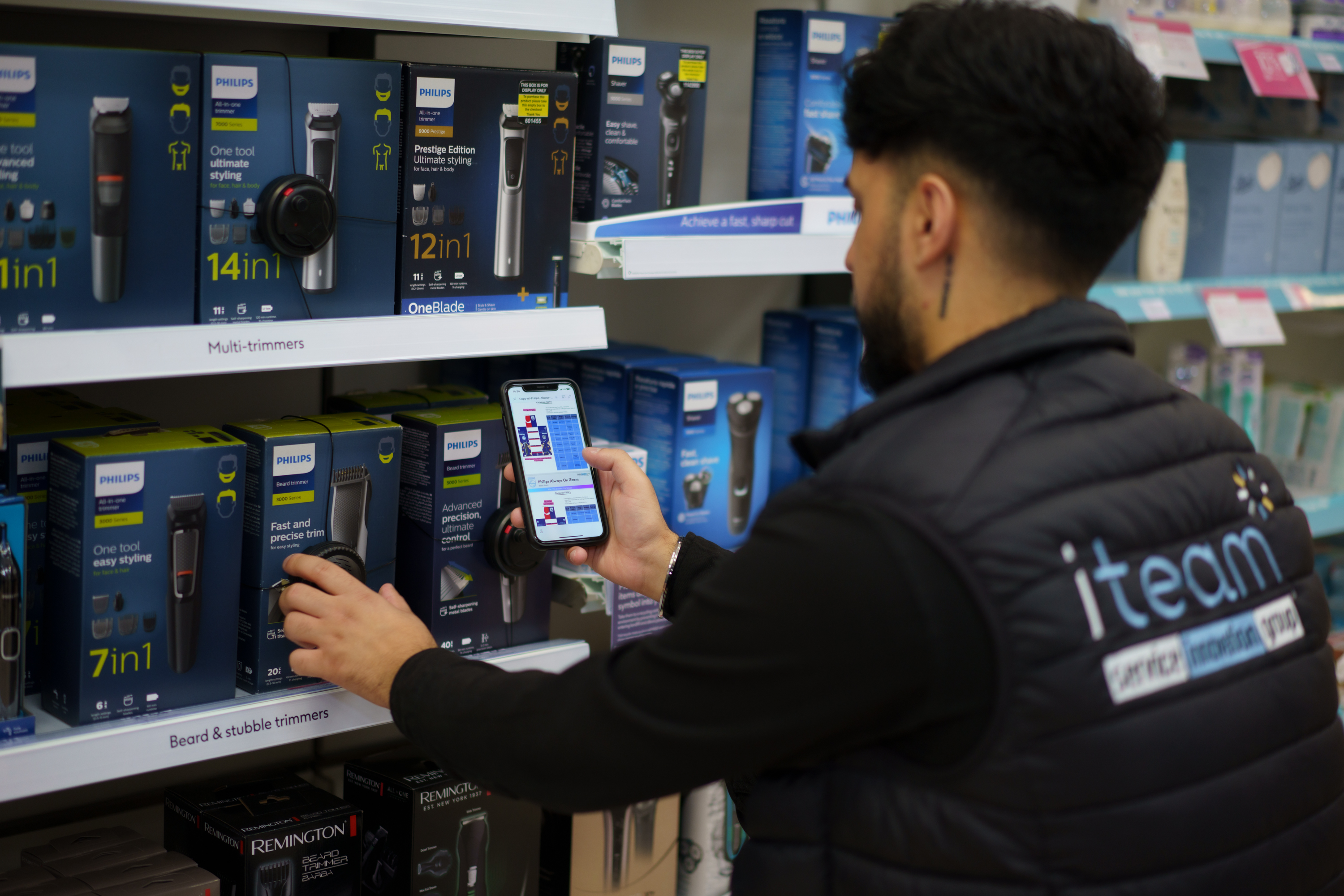 Person in an iteam vest checking Philips beard trimmers on a store shelf while holding a smartphone.