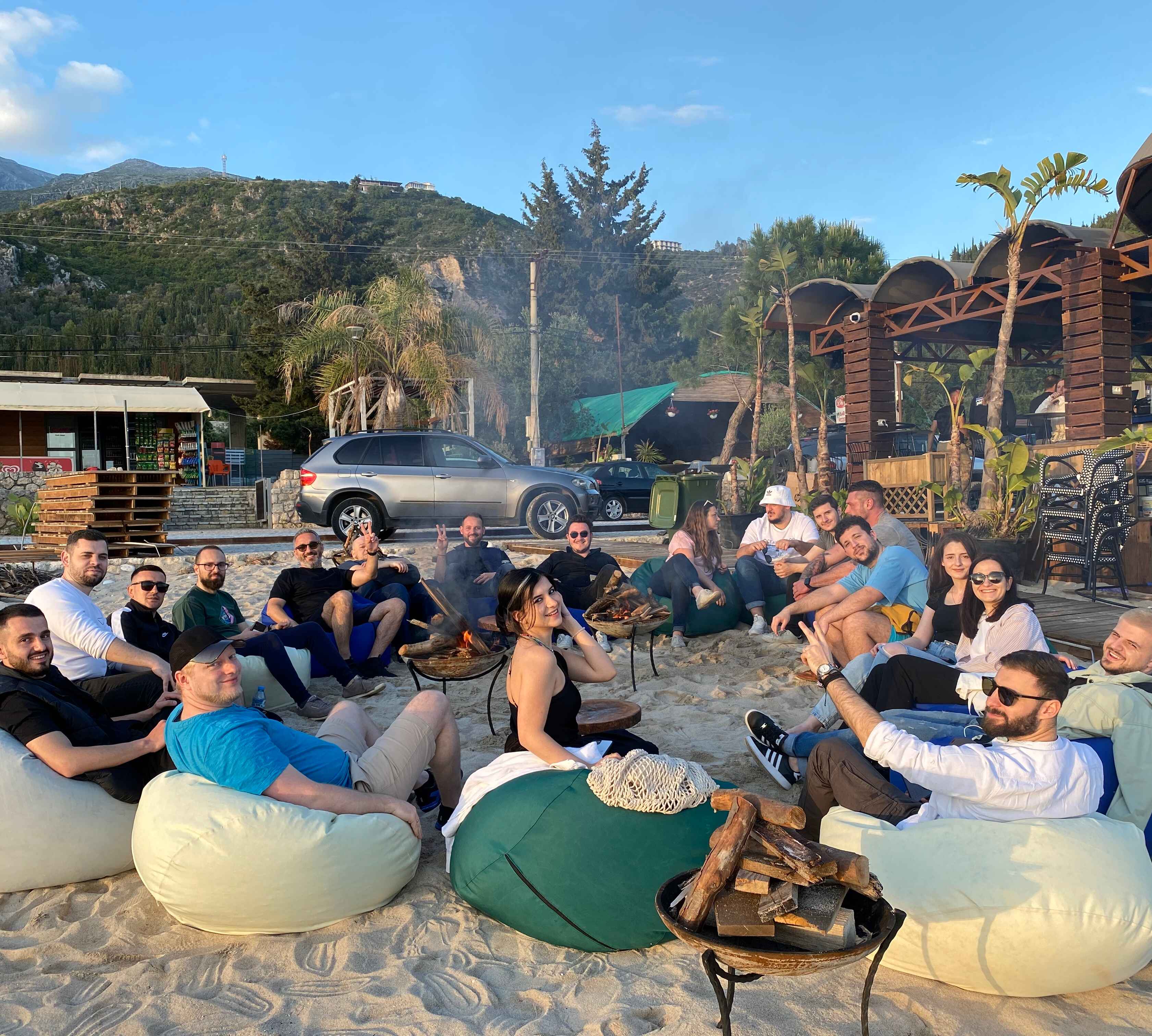 Group of people sitting on bean bags around fire pits on a sandy beach with mountains and palm trees in the background during a sunny day.