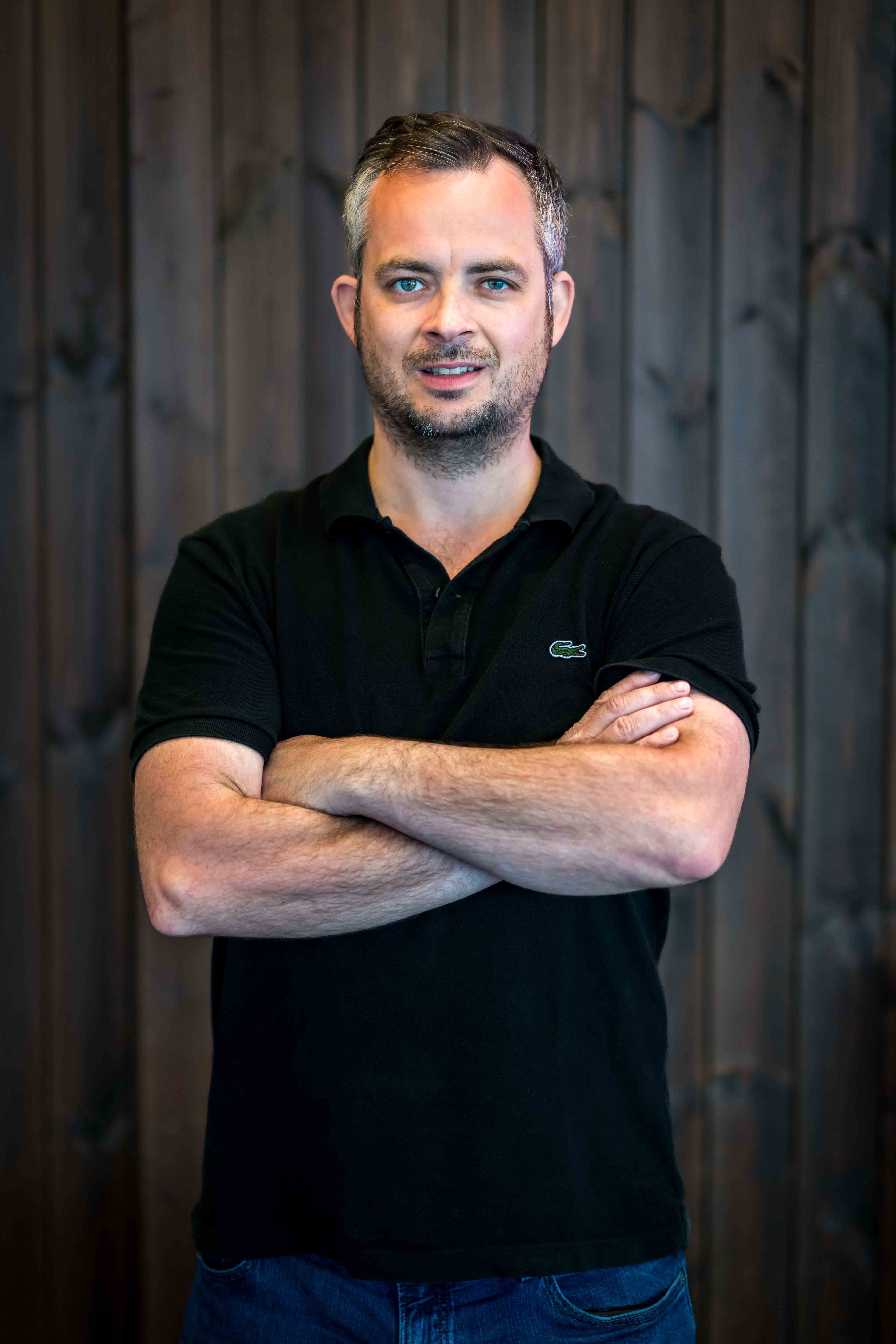 Man with short gray hair and blue eyes wearing a black polo shirt and blue jeans standing with arms crossed in front of a dark wooden background.