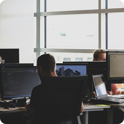 People working at desks with multiple computer monitors in a bright office with large windows.