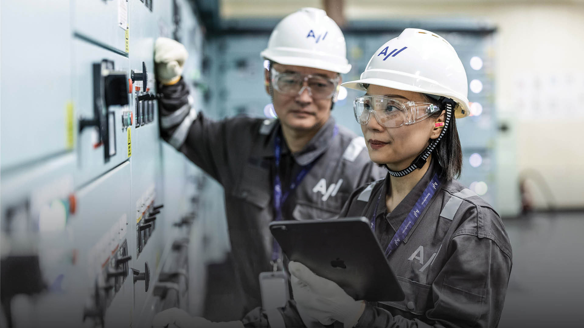 Two industrial workers wearing white helmets and safety glasses, checking control panels with one holding a tablet.
