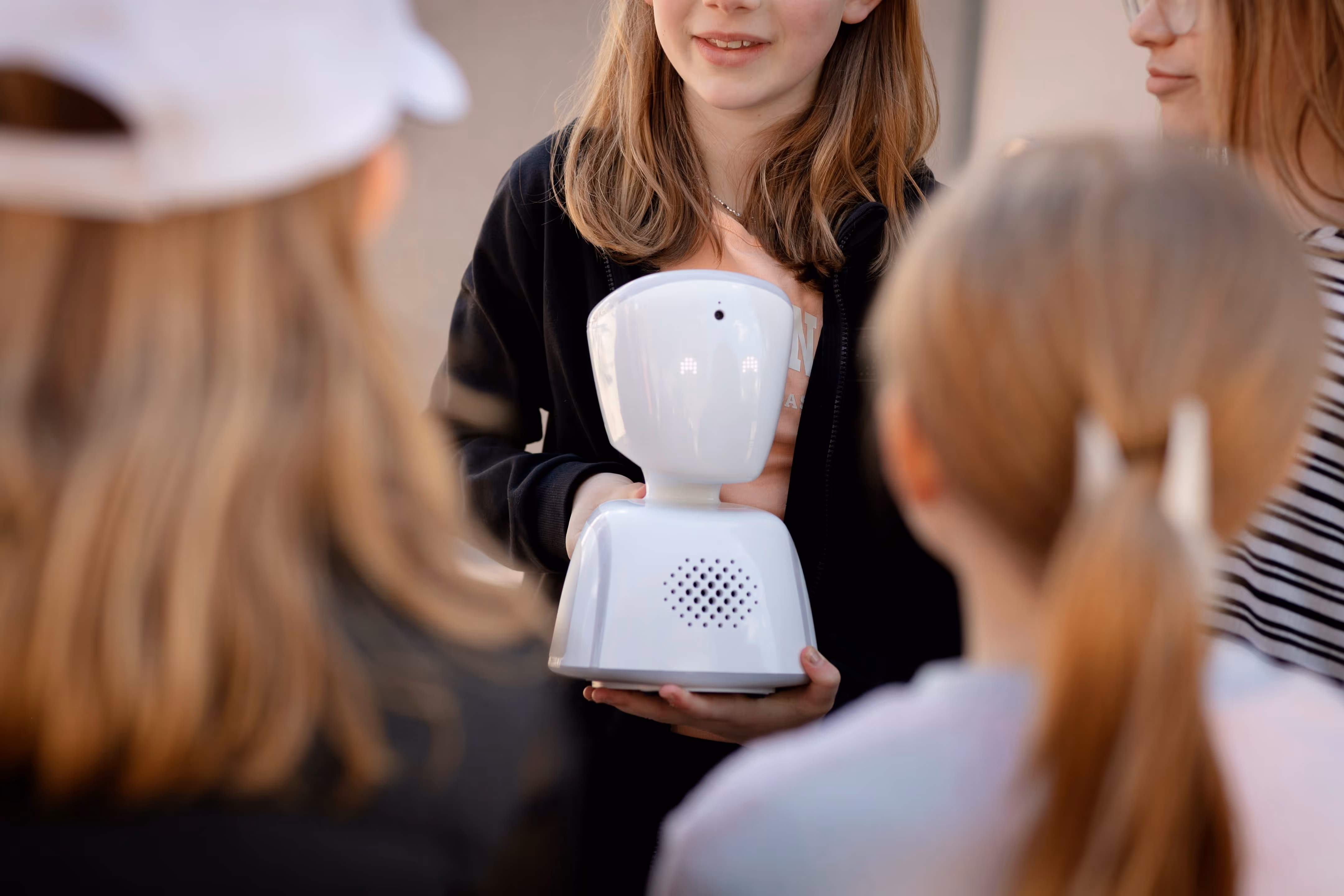 Cuatro niñas sonriendo y sosteniendo un robot AV1. Están afuera, en el patio de un colegio