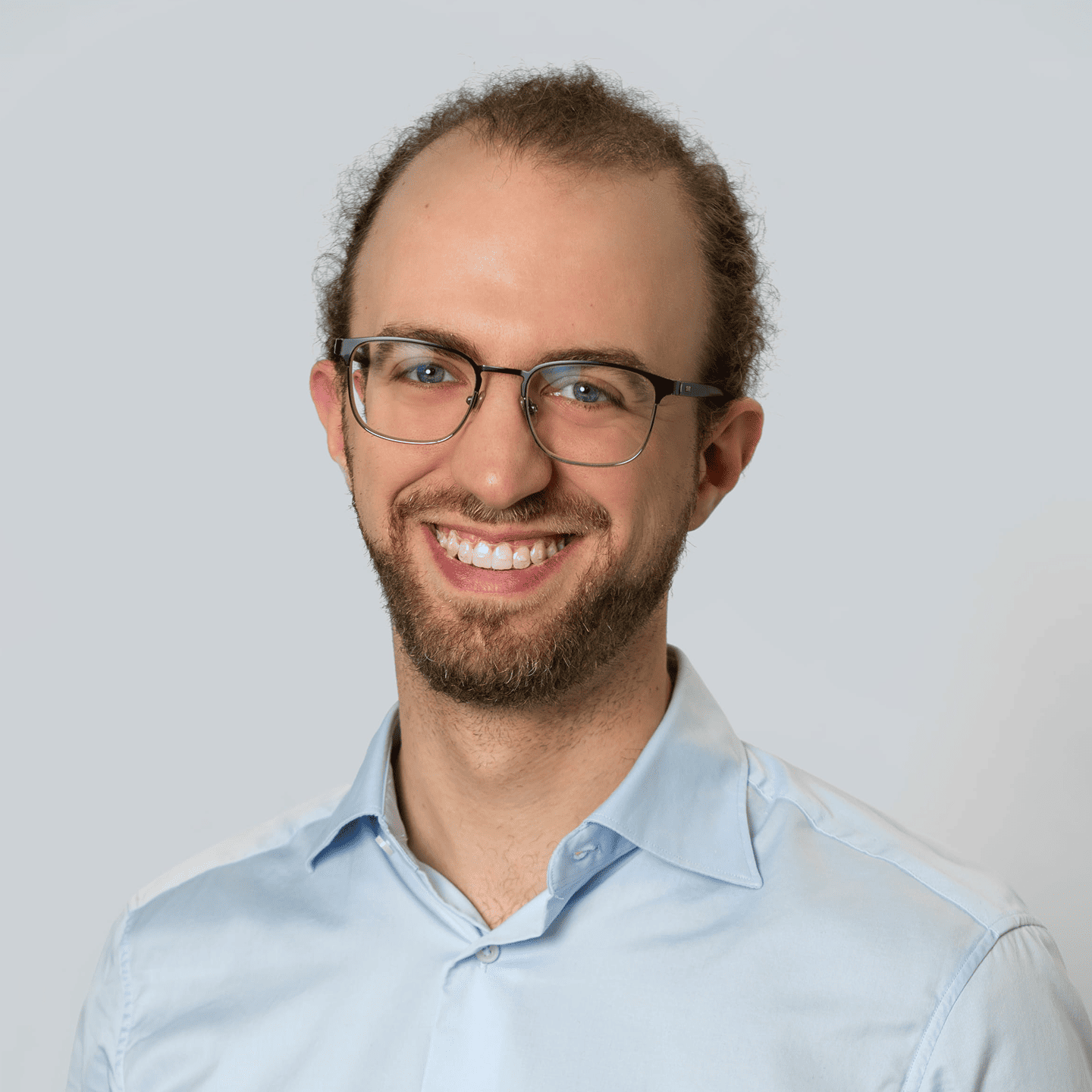 Smiling man with glasses and curly hair wearing a navy blue button-up shirt against a plain light gray background.