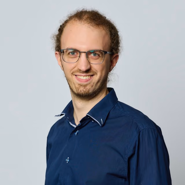 Smiling man with glasses and curly hair wearing a navy blue button-up shirt against a plain light gray background.