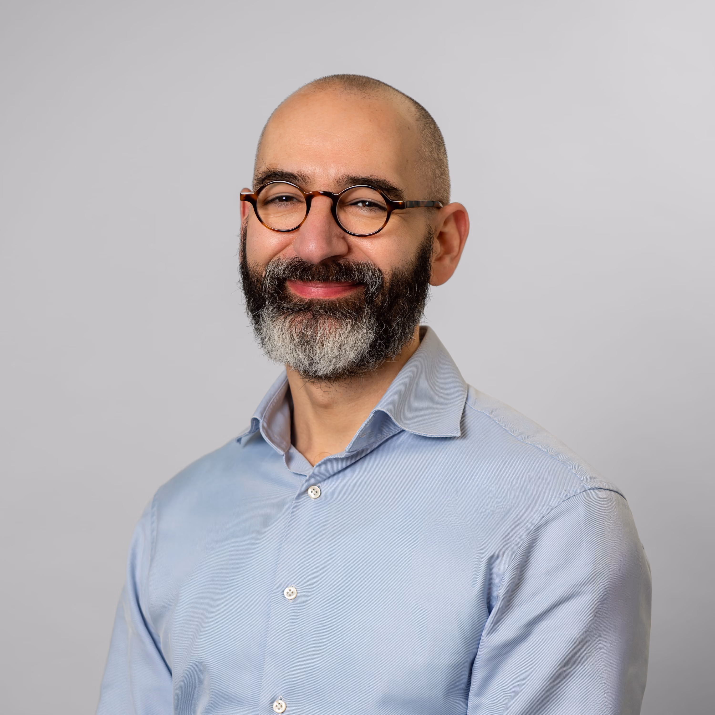 Smiling bald man with a graying beard wearing round glasses and a light blue button-up shirt.