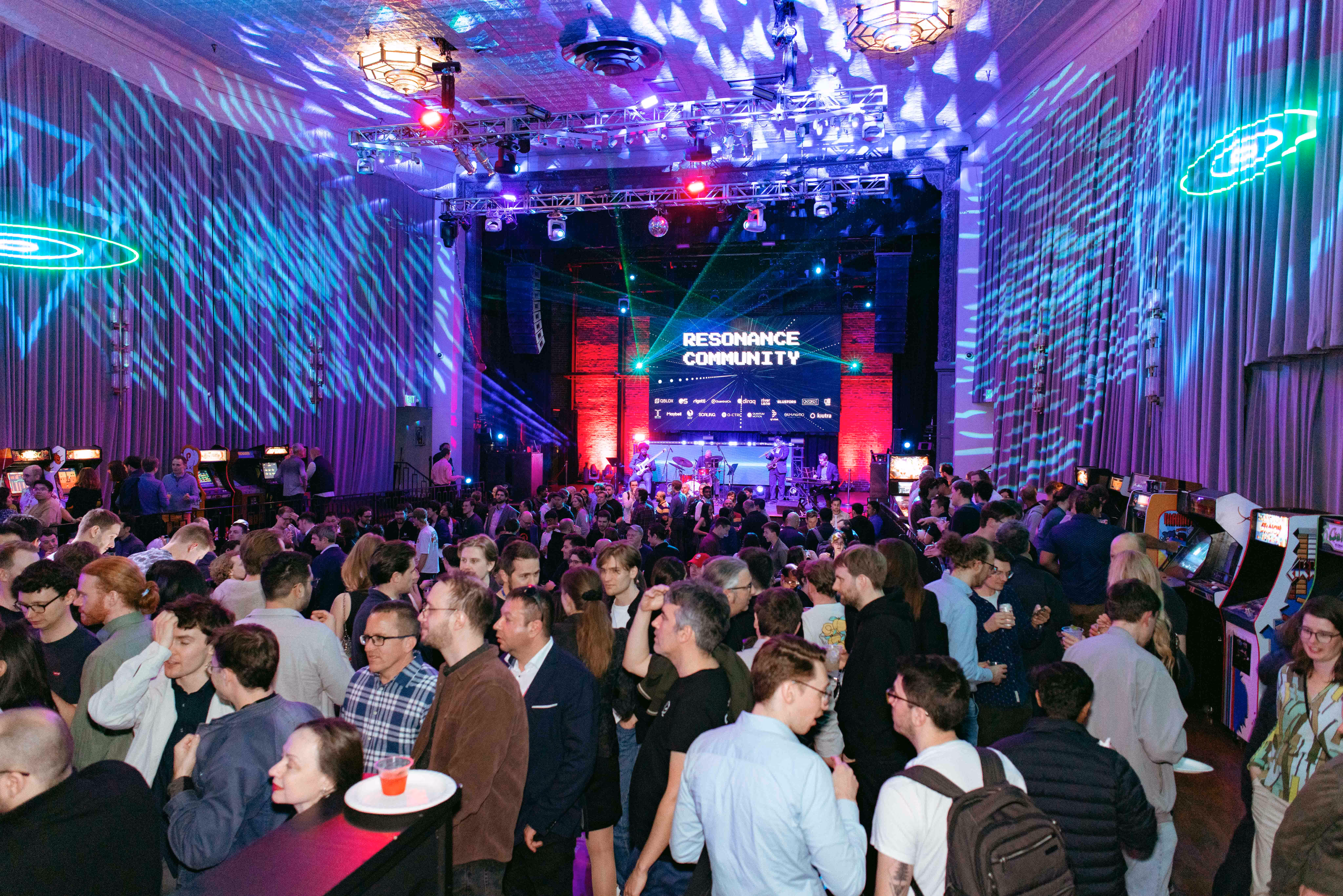 Crowded indoor event with people socializing under colorful stage lights and a screen reading 'Resonance Community.'