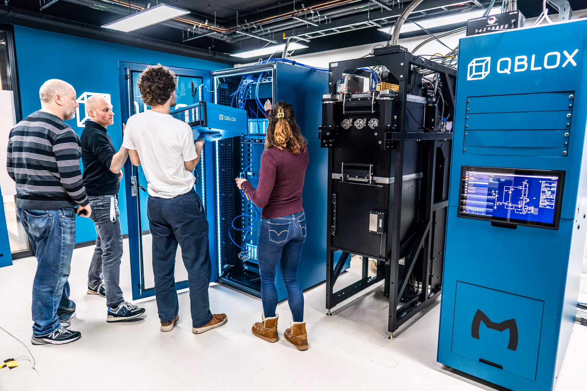 Four people working with a large blue Qblox Cluster rack in a modern lab; one person in white shirt holding a server component, another woman pointing inside the rack, with a Qblox Cluster and screen showing a technical schematic visible.