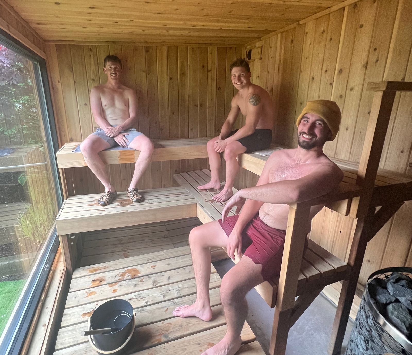 Three shirtless men sitting on wooden benches inside a sauna room with a large window and a bucket with a ladle on the floor.
