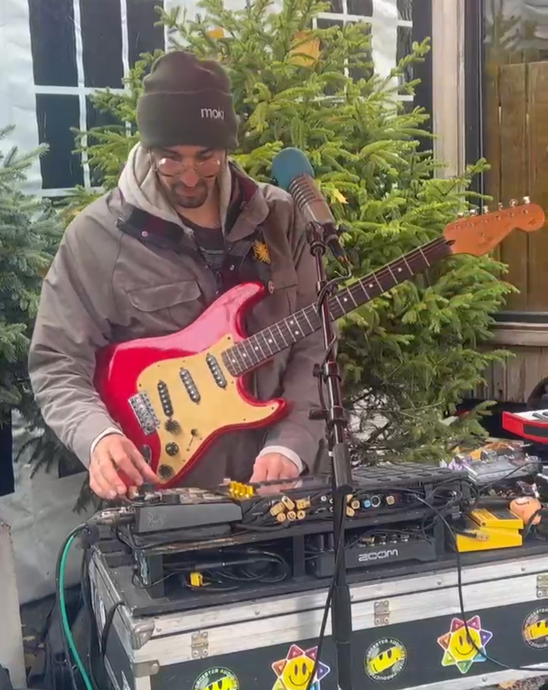 Man wearing glasses and a beanie playing a red electric guitar and adjusting music equipment outdoors near green pine trees.