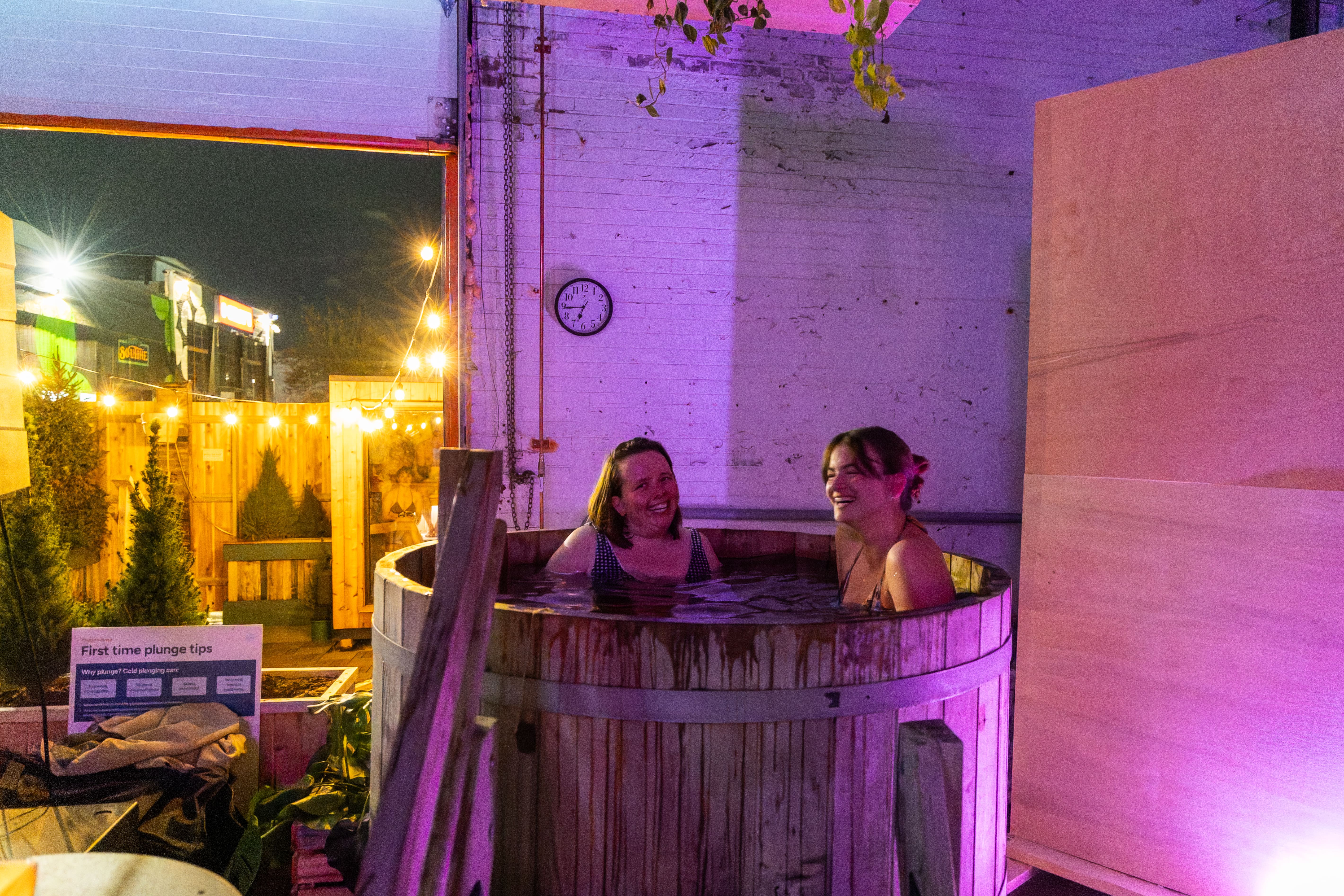 Two women smiling and relaxing in a wooden hot tub at night with string lights and a wall clock in the background.