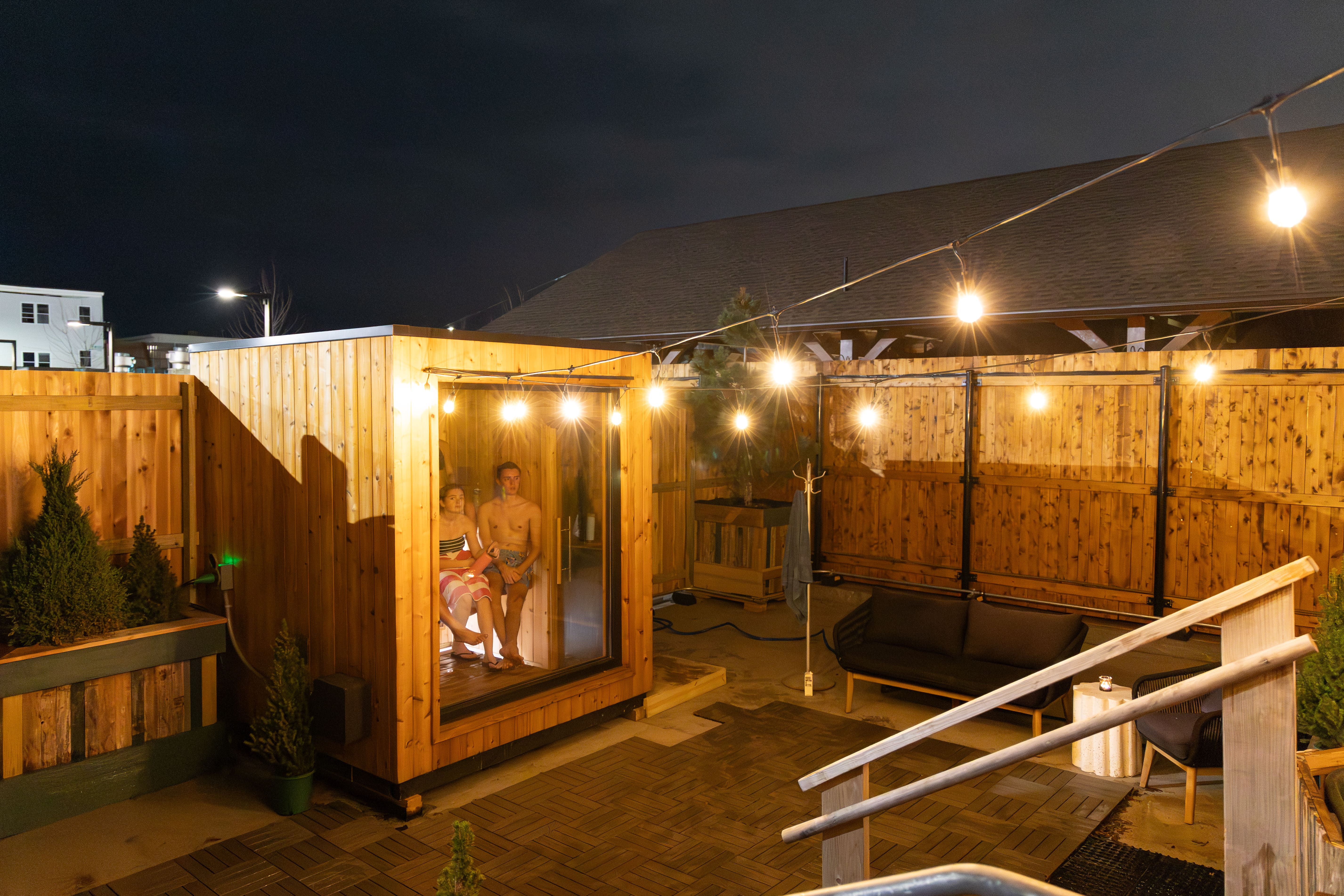 A young couple sitting inside a small wooden sauna illuminated by string lights on a fenced outdoor patio at night.