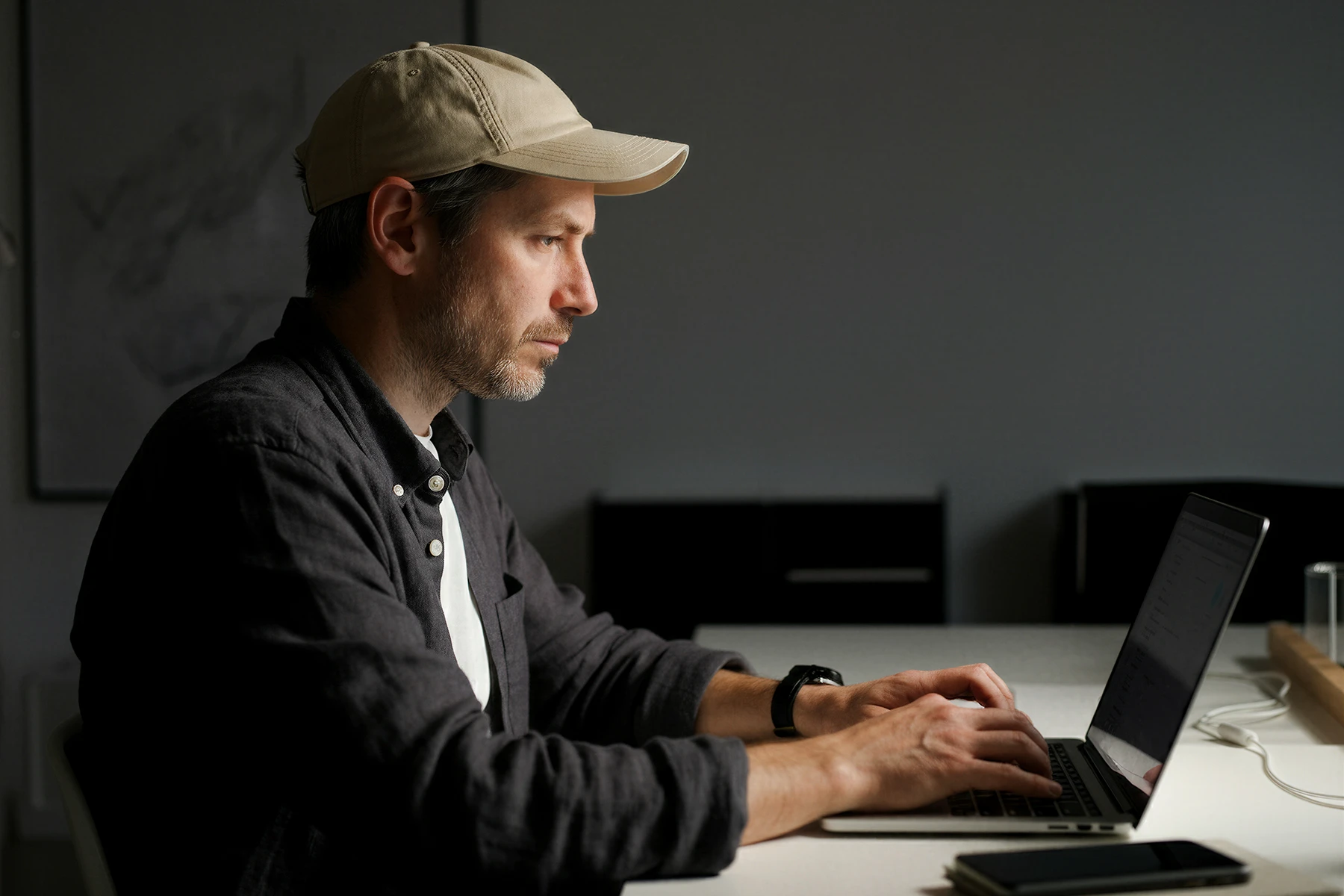 Man wearing a beige cap and dark shirt typing on a laptop at a white desk in a dimly lit room.