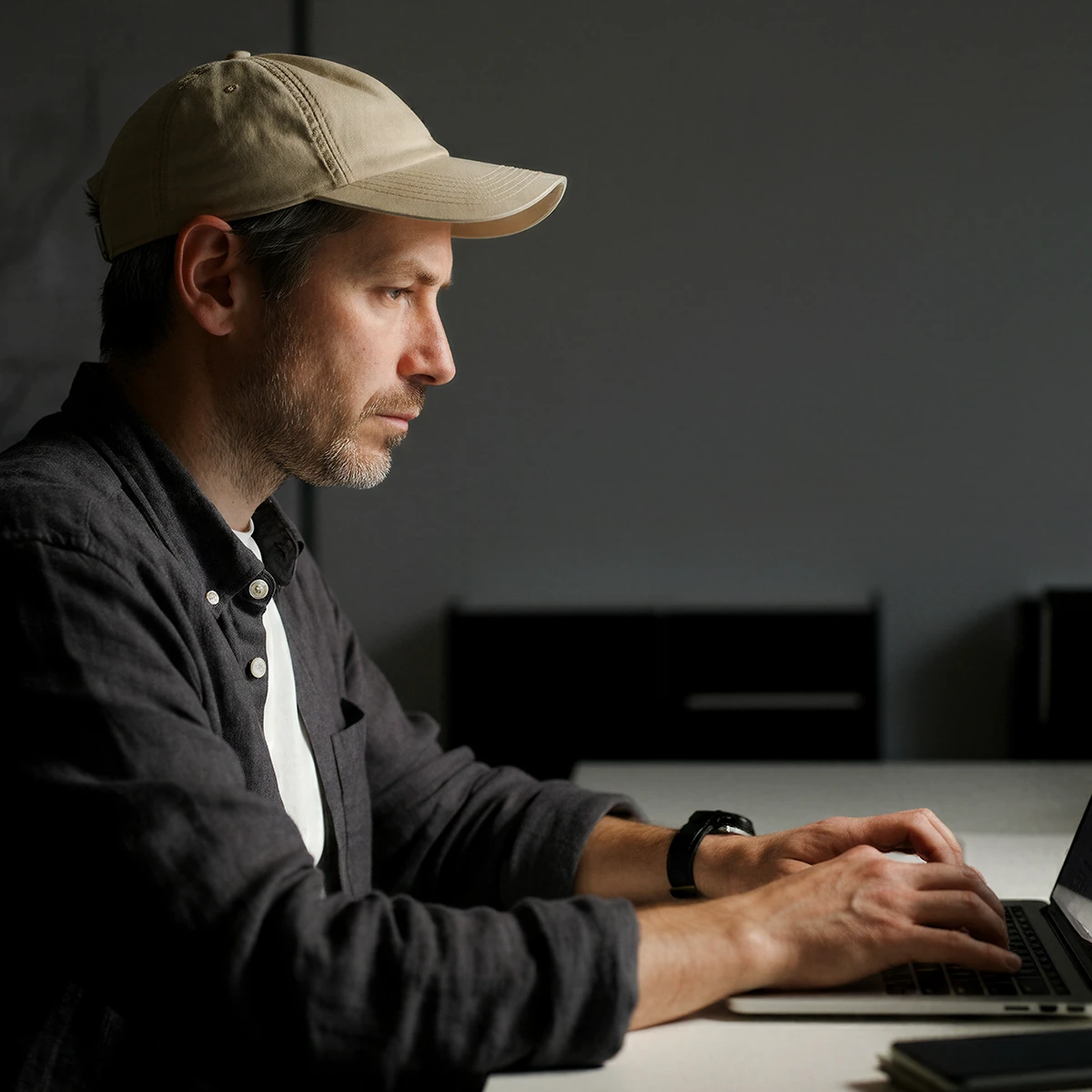 Man wearing a beige cap and dark shirt typing on a laptop at a white table in a dimly lit room.