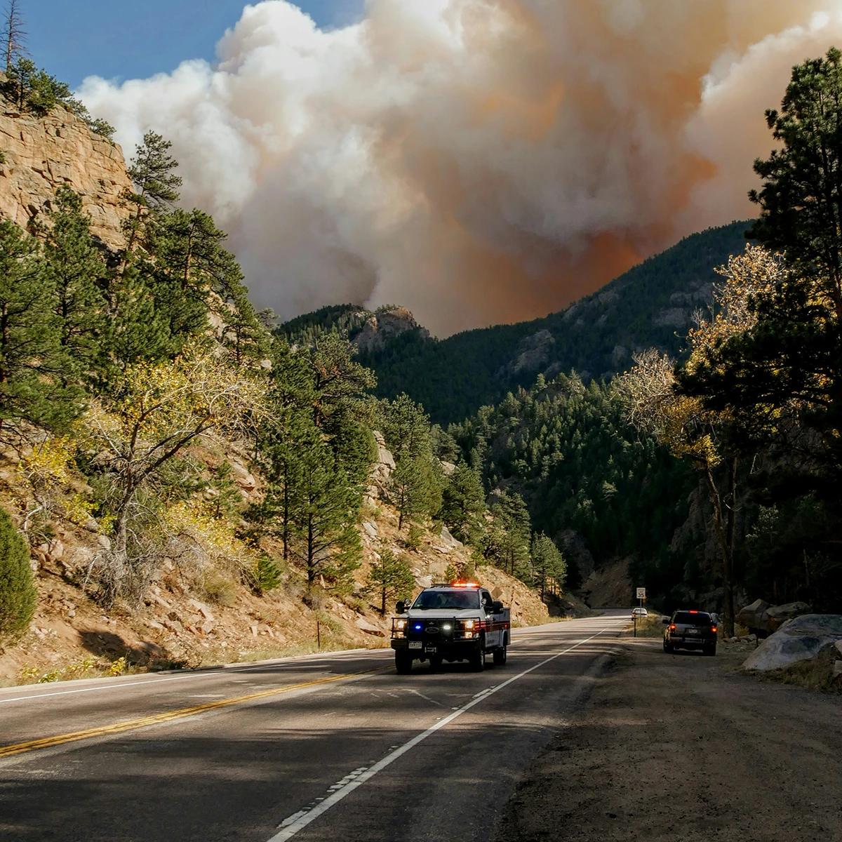 Emergency vehicle with flashing lights driving on a mountain road with heavy smoke from a wildfire in the background.
