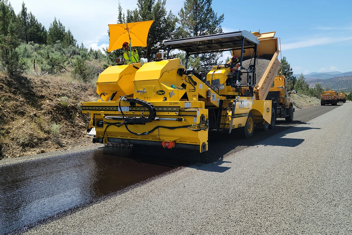 Yellow road paving machine laying asphalt on a rural road with workers operating it under a clear sky.