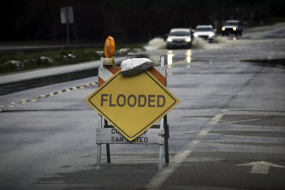 Yellow flooded road sign on wet street with cars driving through floodwater in the background.