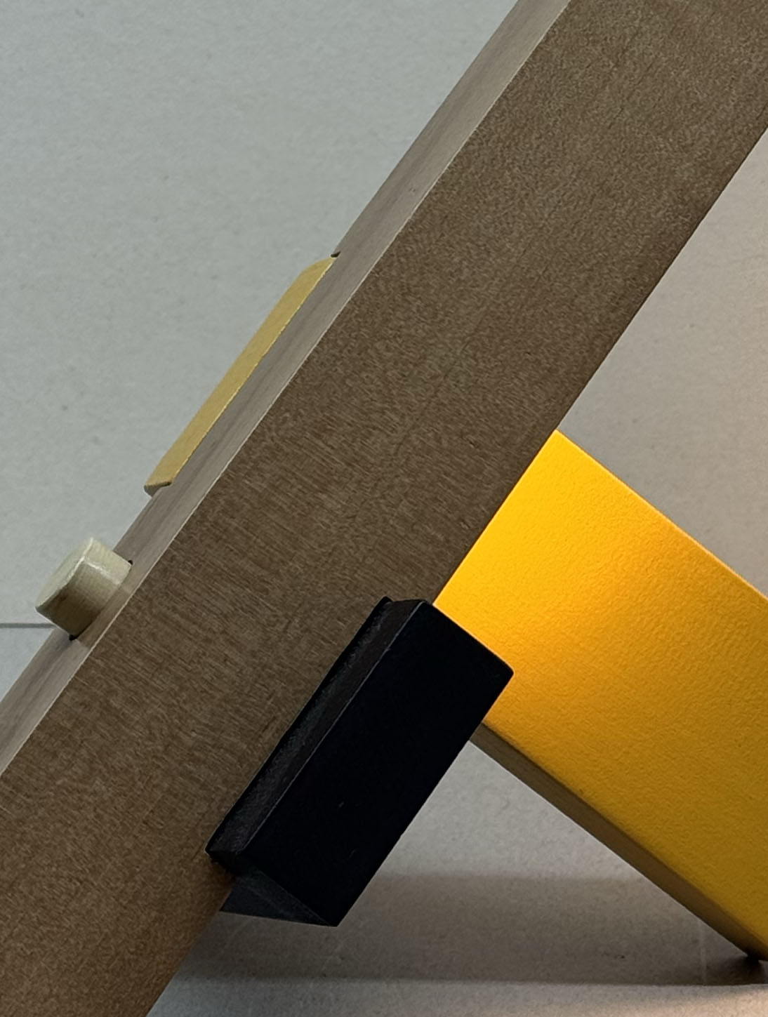 Close-up of a wooden structure with a black triangular piece attached, next to a yellow panel, set against a neutral background.