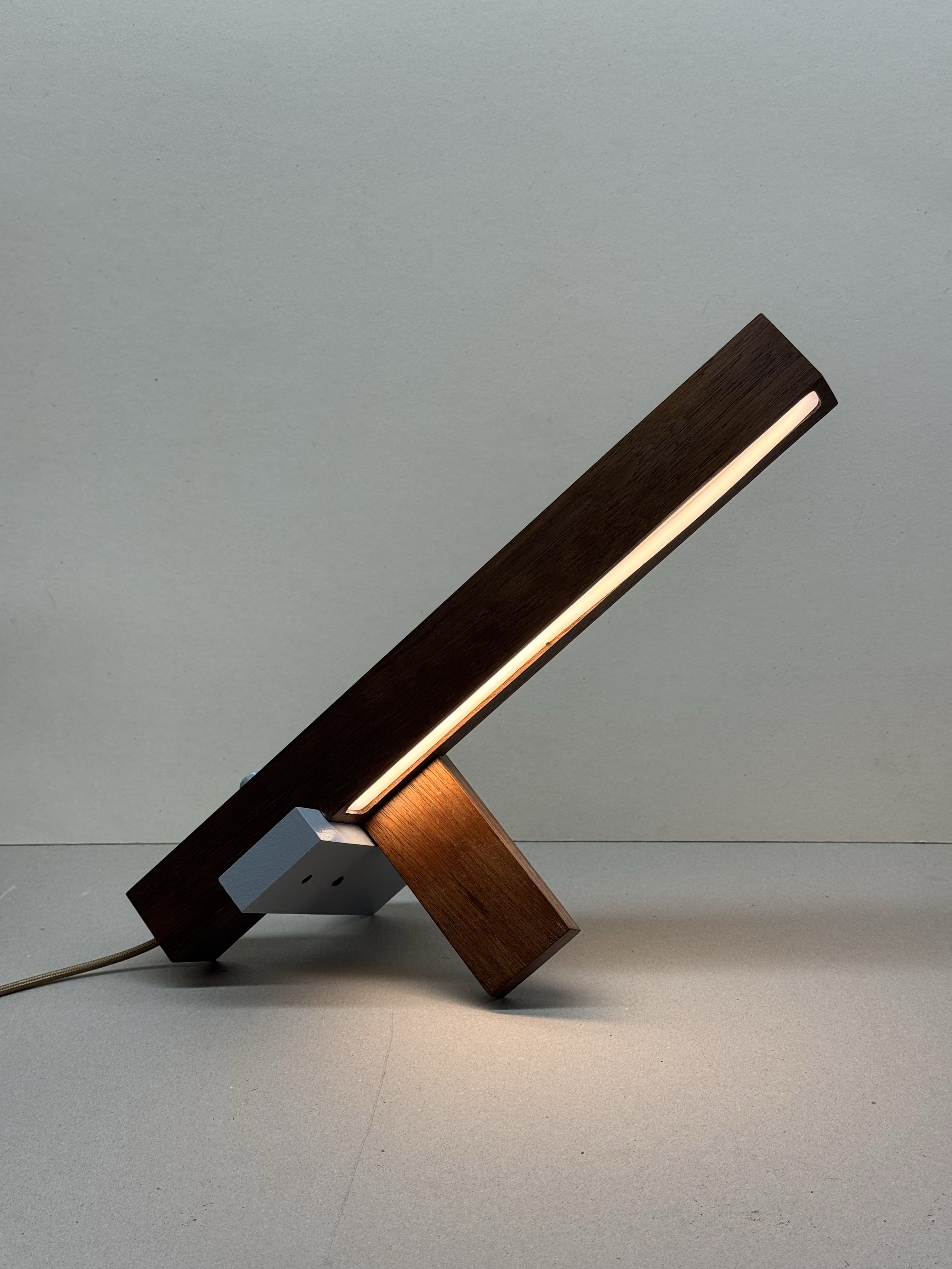Modern wooden desk lamp with a rectangular light strip, supported by a smaller angled wooden block and a gray metal piece on a flat surface.